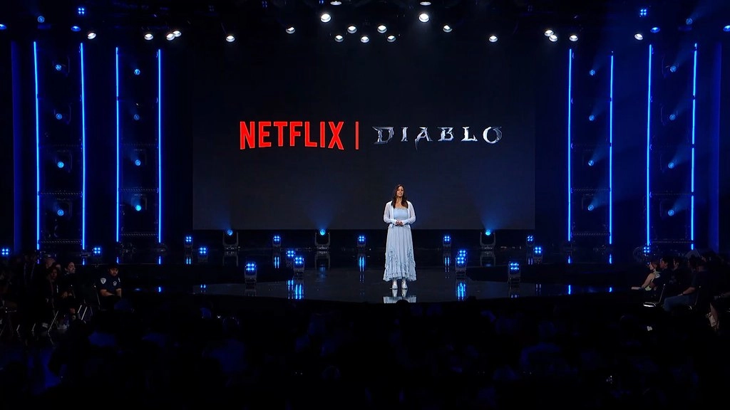 Johanna Faries w jasnej sukni stoi na oświetlonej na niebiesko scenie BlizzConu przed ekranem z logotypami Netflixa i Diablo. Na pierwszym planie widać sylwetki widzów. Źródło: transmisja BlizzConu na kanale Blizzarda na Twitchu.